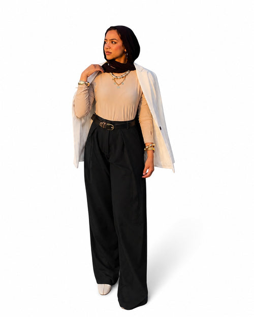A woman modeling a stylish outfit consisting of a beige ribbed long-sleeve top, black wide-leg trousers with a black belt, a white open-front blazer draped over her shoulders, and a black headscarf, set against a plain white background.