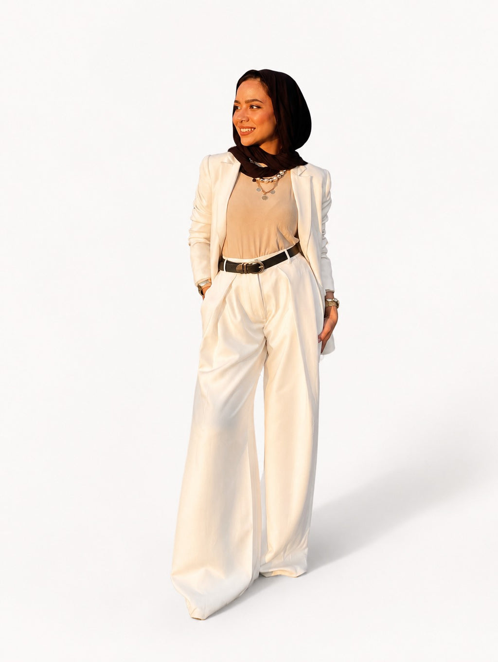 A woman modeling a white wide-leg pantsuit with a beige top, black belt, and black scarf, standing against a plain white studio background.