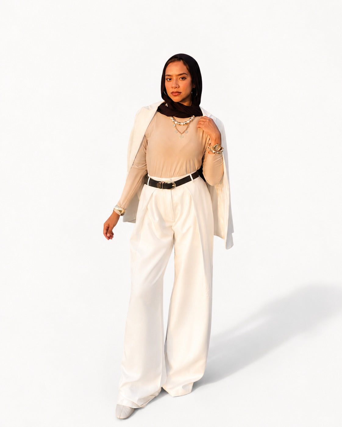 A model wearing a beige long-sleeve top, white wide-leg trousers, a white draped cape-style jacket, a black belt, and a black headscarf, standing against a plain white background.