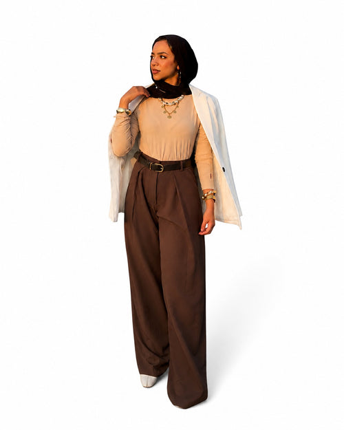 A woman modeling a stylish outfit consisting of a beige long-sleeve top, dark brown wide-leg trousers with a dark brown belt, a black hijab, gold necklaces, and a white open-front blazer, set against a plain white background.